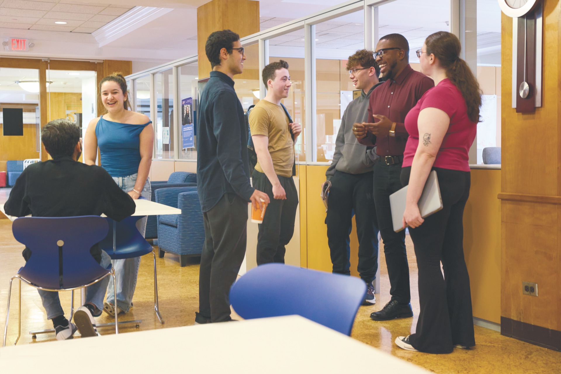 Students gather in a lounge, laughing and talking in small groups around tables and blue chairs.