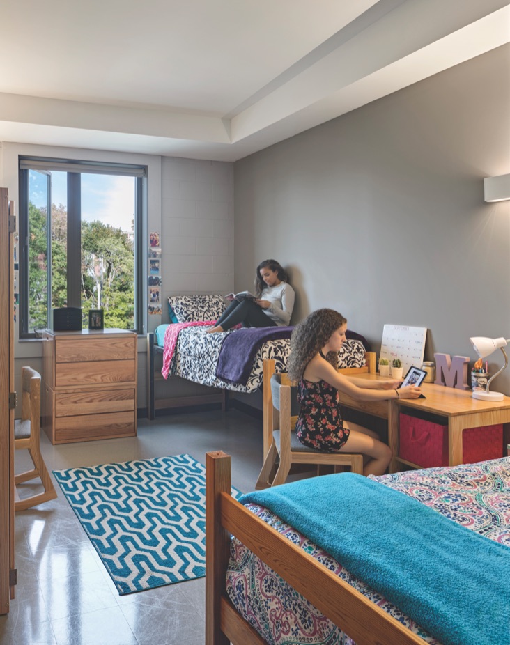 A student reading on her bed in a residence hall room decorated with patterned bedding and string lights.