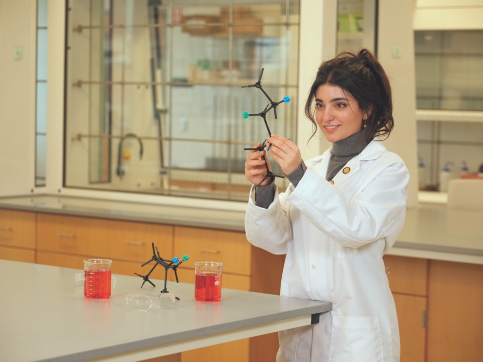 A Framingham State student in a white lab coat holds a molecular model in a chemistry laboratory.
