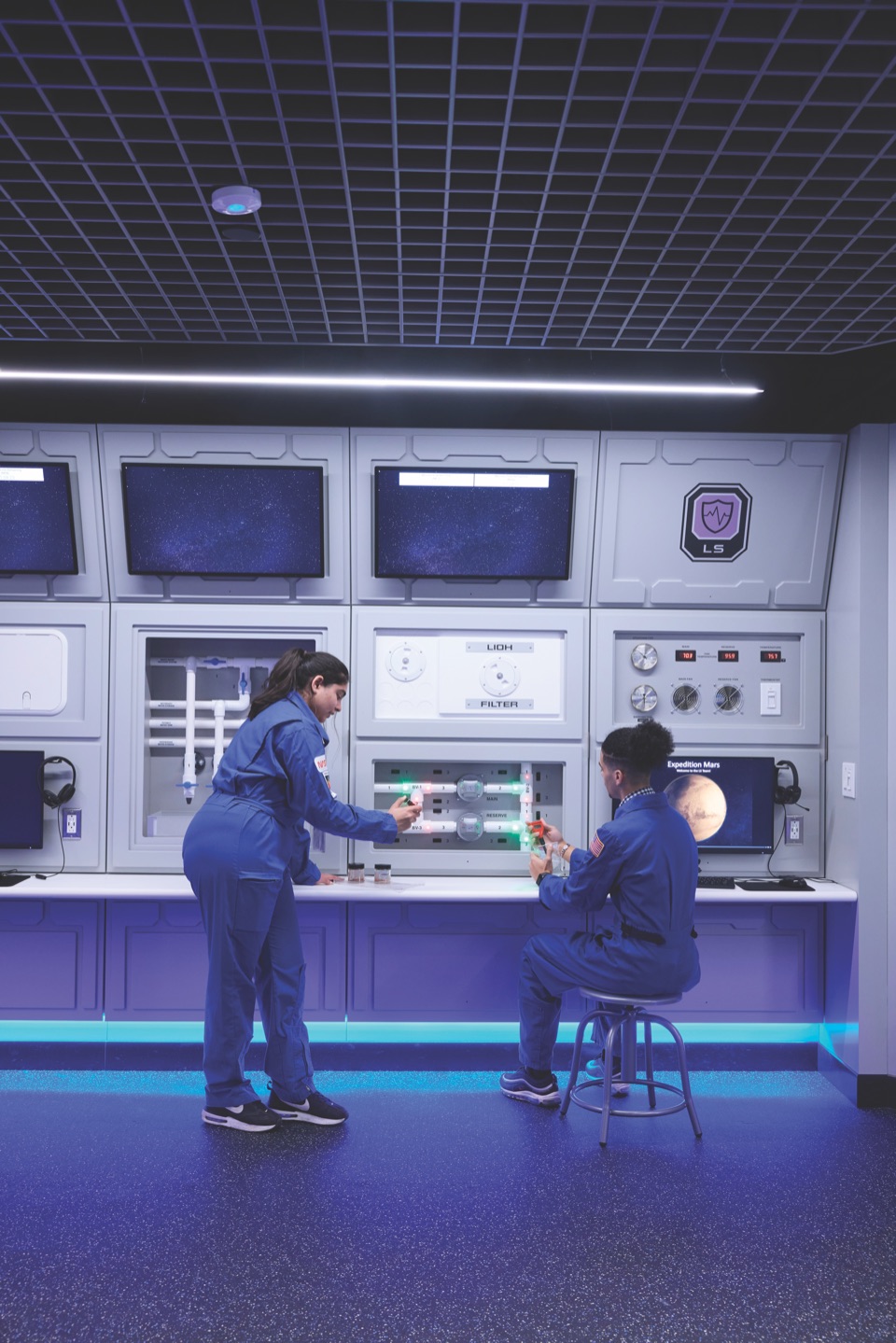 Two students in blue coveralls operating an instrument at a lab bench at the Christa McAuliffe Center.