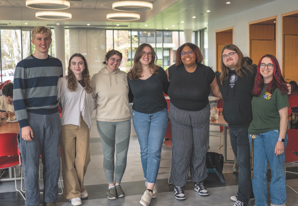 Seven Framingham State students standing arm in arm in a residence-hall lounge, smiling for the camera.