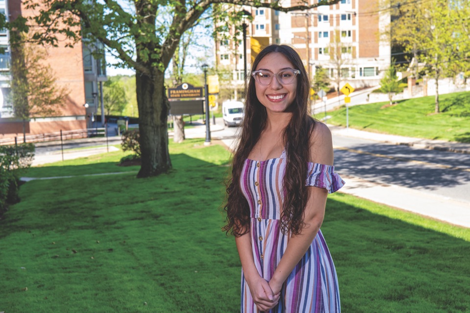 Bianca Furtado standing on a campus lawn, residence halls and the Framingham State entrance sign behind her.