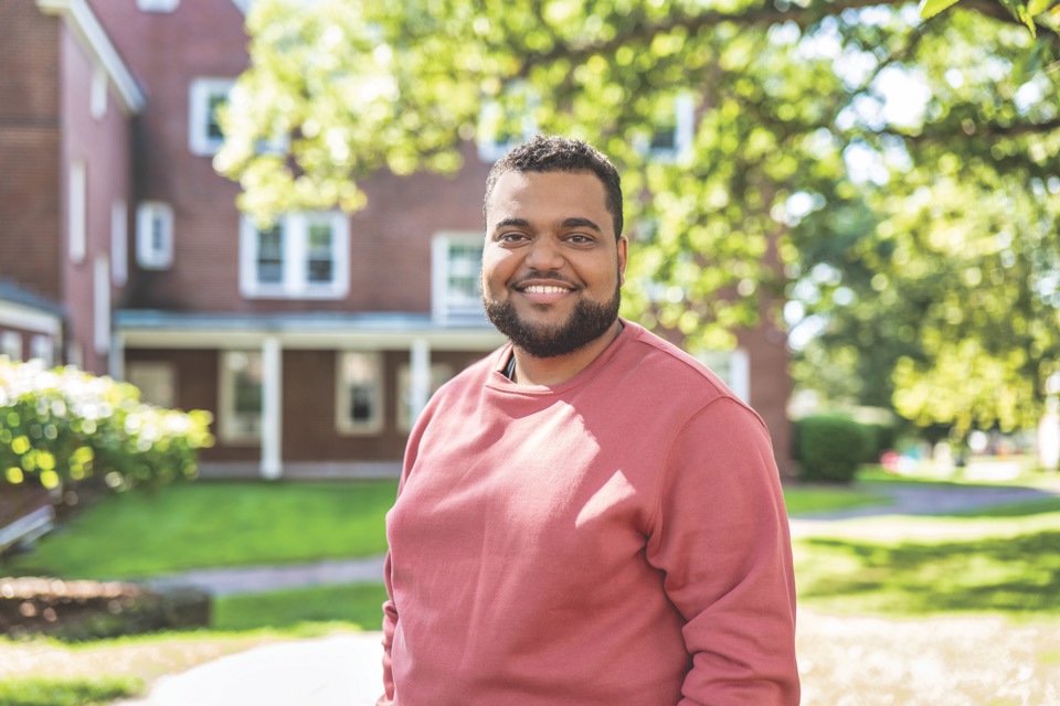 Cesar Matos smiling outdoors on campus in a rose-coloured sweatshirt, with a brick residence hall and summer trees behind him.