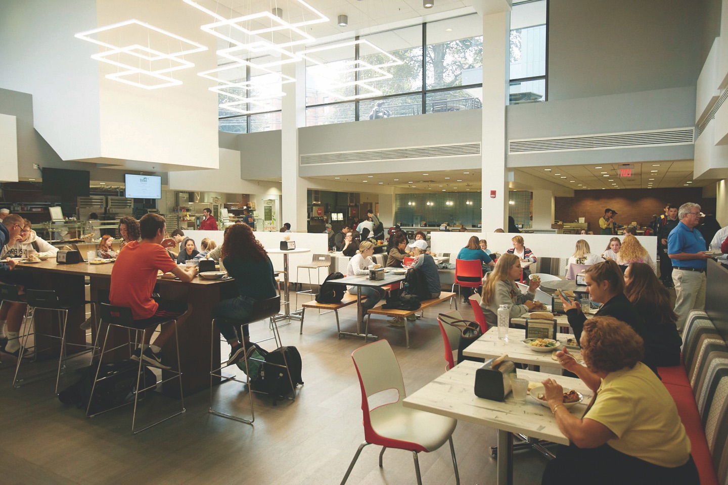 Students working and talking together at high tables in a bright, double-height campus centre lounge with a wall of windows.