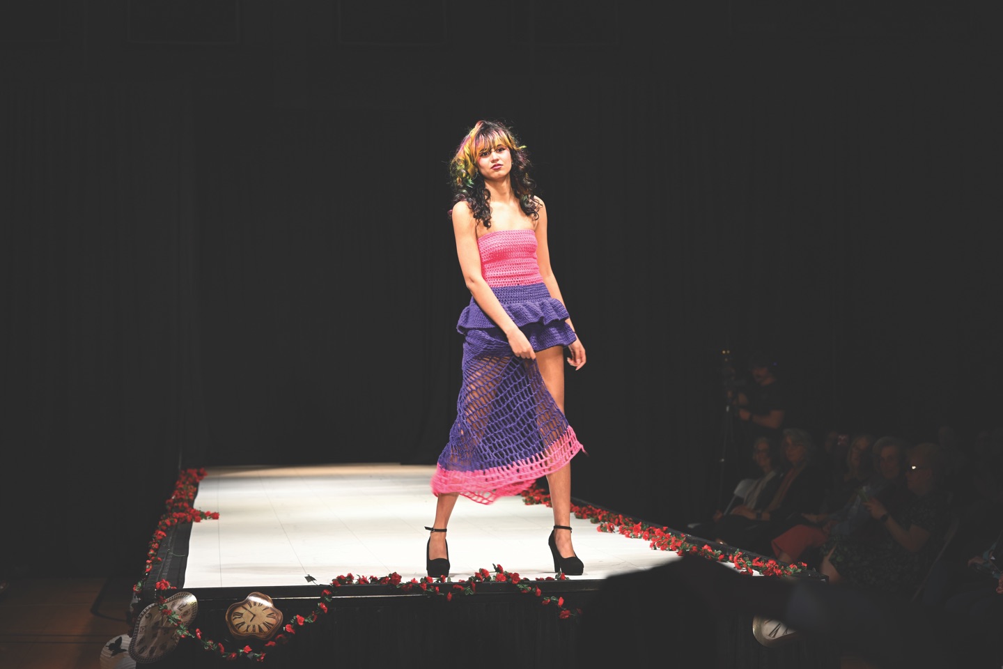 A student walking a runway in a handmade knitted dress at the Student Fashion Show, along a floor scattered with rose petals.