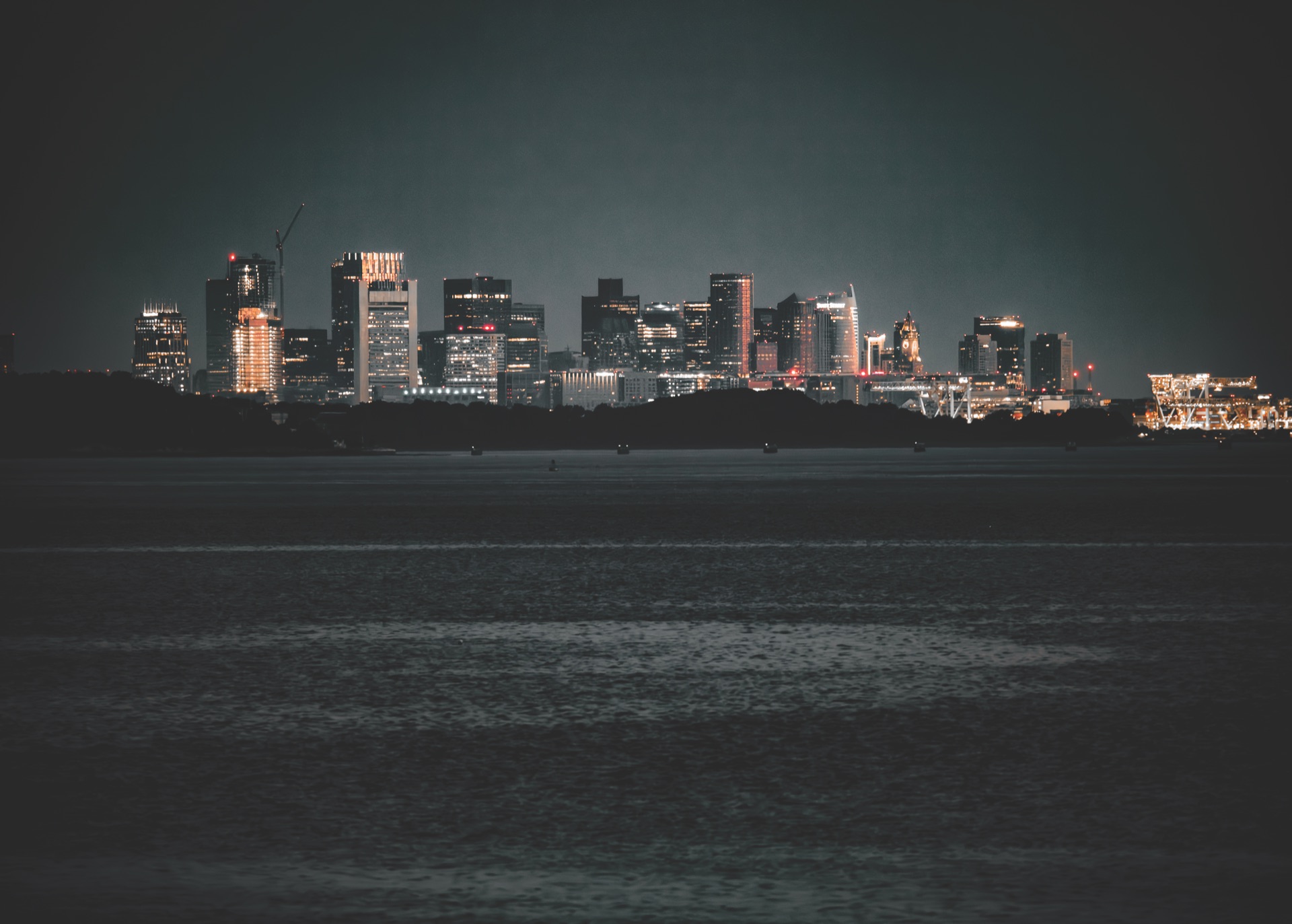 The Boston skyline at night, its office towers lit gold and white against a dark sky and reflected in the harbour water.