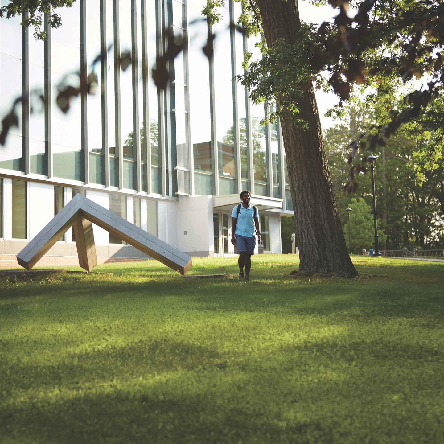 A student with a backpack walking across a sunlit campus lawn, a modern glass-fronted academic building and a large shade tree behind them.