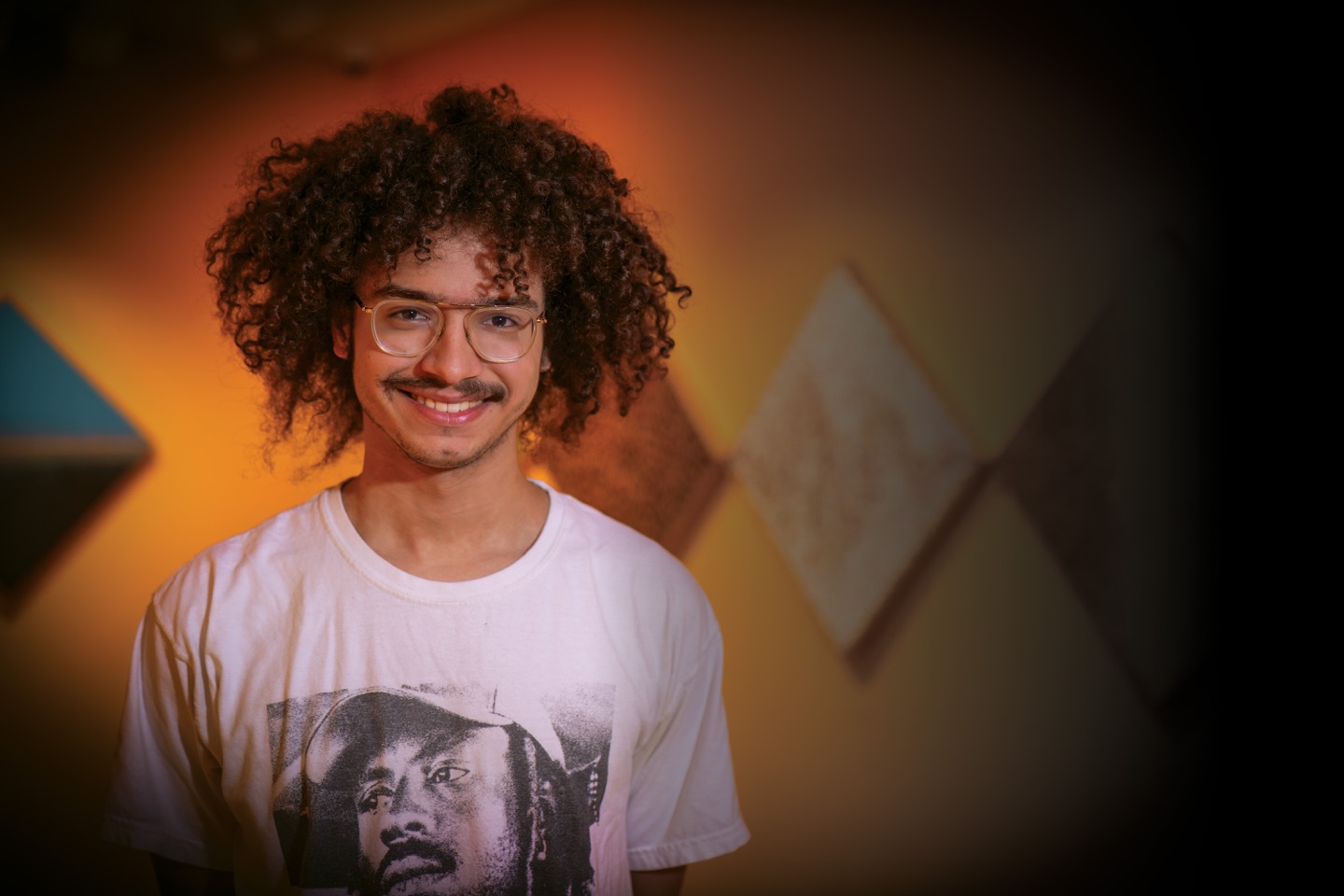 A student with curly hair, glasses and a mustache smiling at the camera.